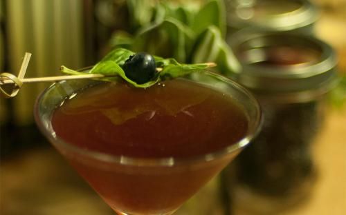  Skewered black olive on a toothpick placed on the glass rim of a basil-infused pomegranate martini drink